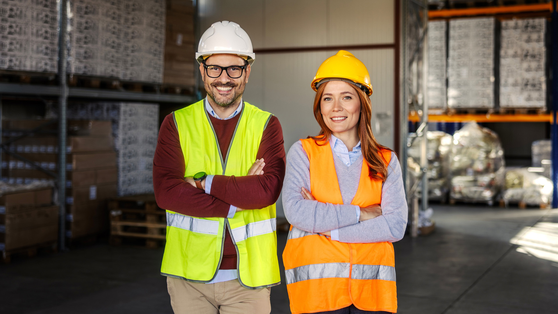 Två personer i skyddshjälmar och reflexvästar står med armarna i kors i ett lager, med hyllor och pallar i bakgrunden.