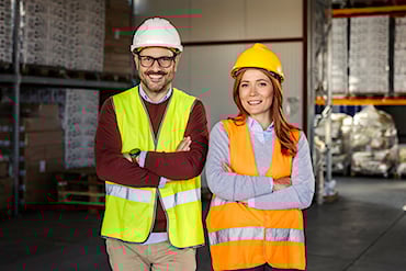 Två personer i skyddshjälmar och reflexvästar står med armarna i kors i ett lager, med hyllor och pallar i bakgrunden.